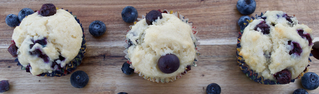 Three blueberry muffins in paper liners on a wooden surface surrounded by fresh blueberries