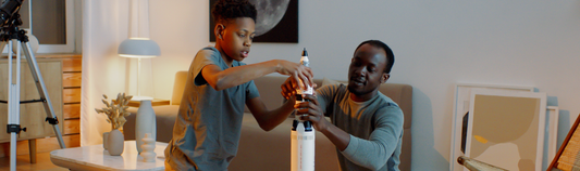 Father and son assembling a model rocket together in a living room, with a telescope nearby