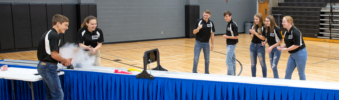 Students in matching black polos watching a CO2 dragster race on a track in a gym as a puff of CO2 releases