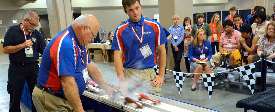 Two men in matching red, white and blue shirts operating a CO2 dragster track while spectators watch behind checkered flags