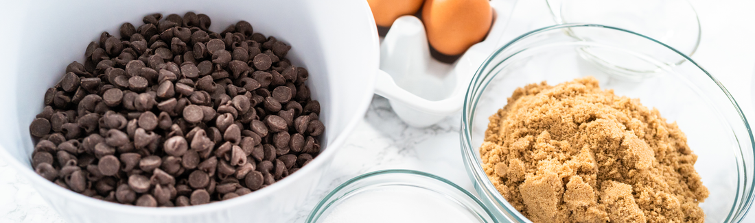 Bowl of chocolate chips beside a bowl of brown sugar and eggs on a white counter