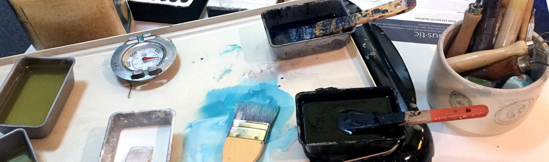 Encaustic painting workspace with wax pans, a brush, a thermometer and tools in a cup