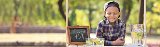 Smiling boy in a cap at a lemonade stand with a chalkboard sign reading 10 cents and a pitcher of lemonade