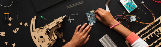 Hands assembling an Arduino circuit board with wires, a breadboard and wooden parts on a black table