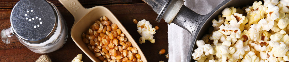 Scoop of popcorn kernels beside a salt shaker and a pot of popped popcorn on a wooden table