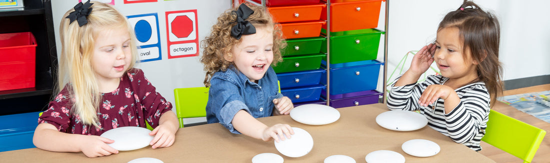 Three preschool-age children laughing at a table with white paper plates in a classroom