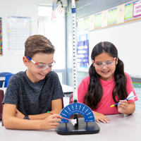 Two elementary-aged students in a classroom setting with safety goggles, one using the straw rocket launcher on a table.