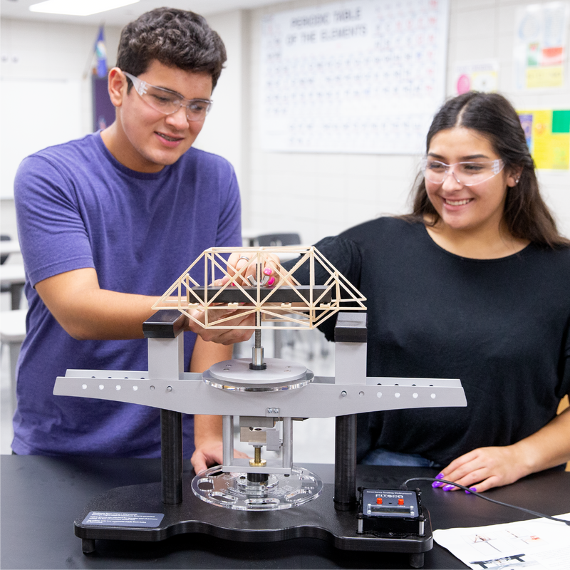 Two students in a classroom setting working on a STEM project testing their bridge with Pitsco Education's structure tester.