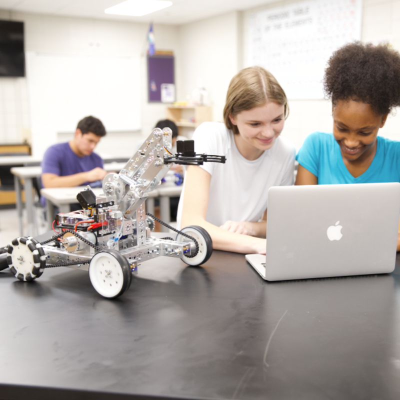 Two students working on a robot and using a laptop in a classroom setting.